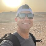 White male standing the desert. He is wearing a ball cap and sunglasses.